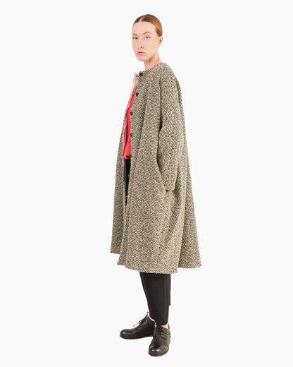 A model is shown in profile wearing a long, open-front coat with a textured beige and gray pattern, layered over a pink top, black pants, and black leather shoes, set against a plain light gray background.